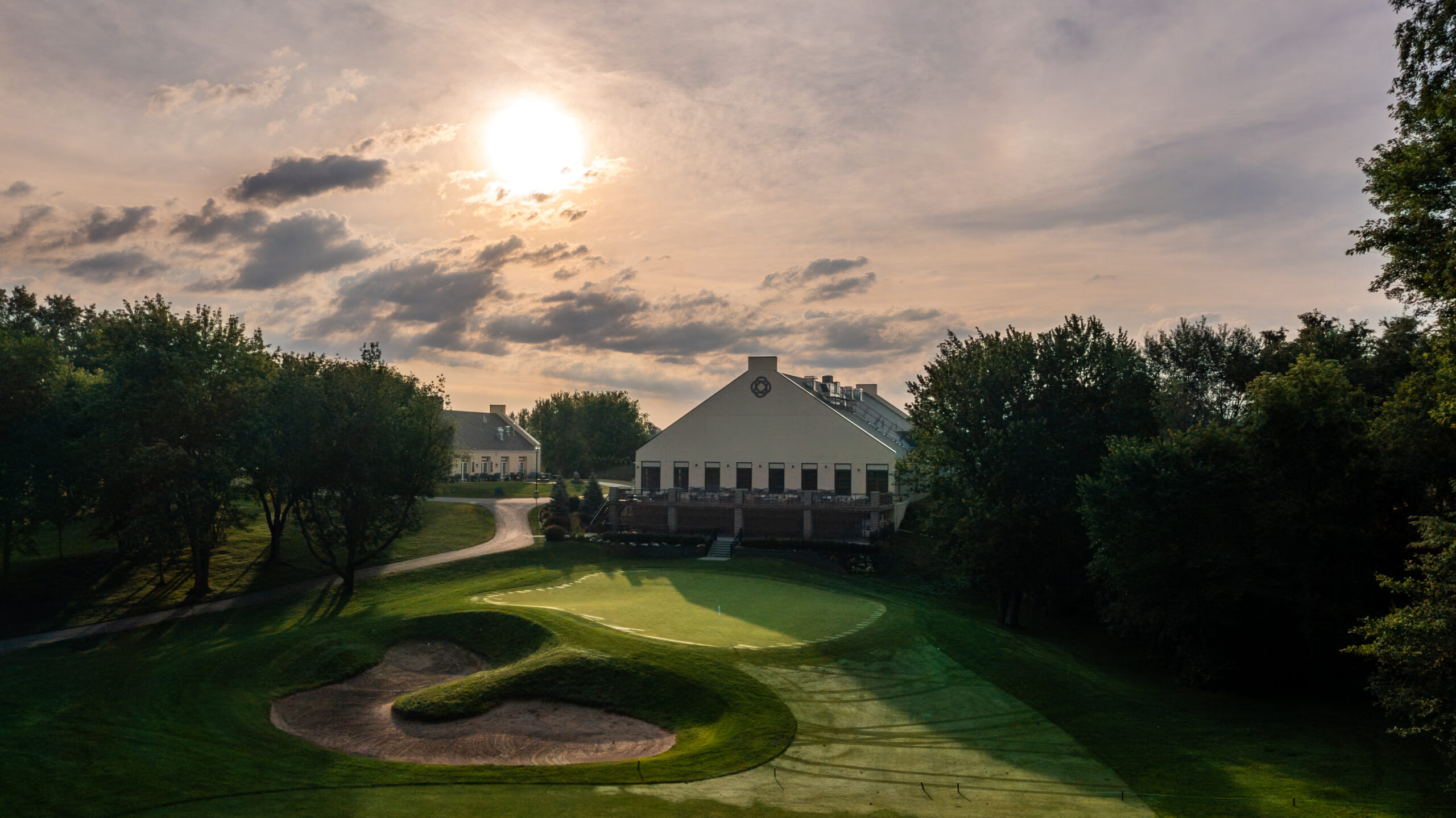 A golf course with a green and sand trap, featuring a clubhouse under a cloudy sky at sunset.